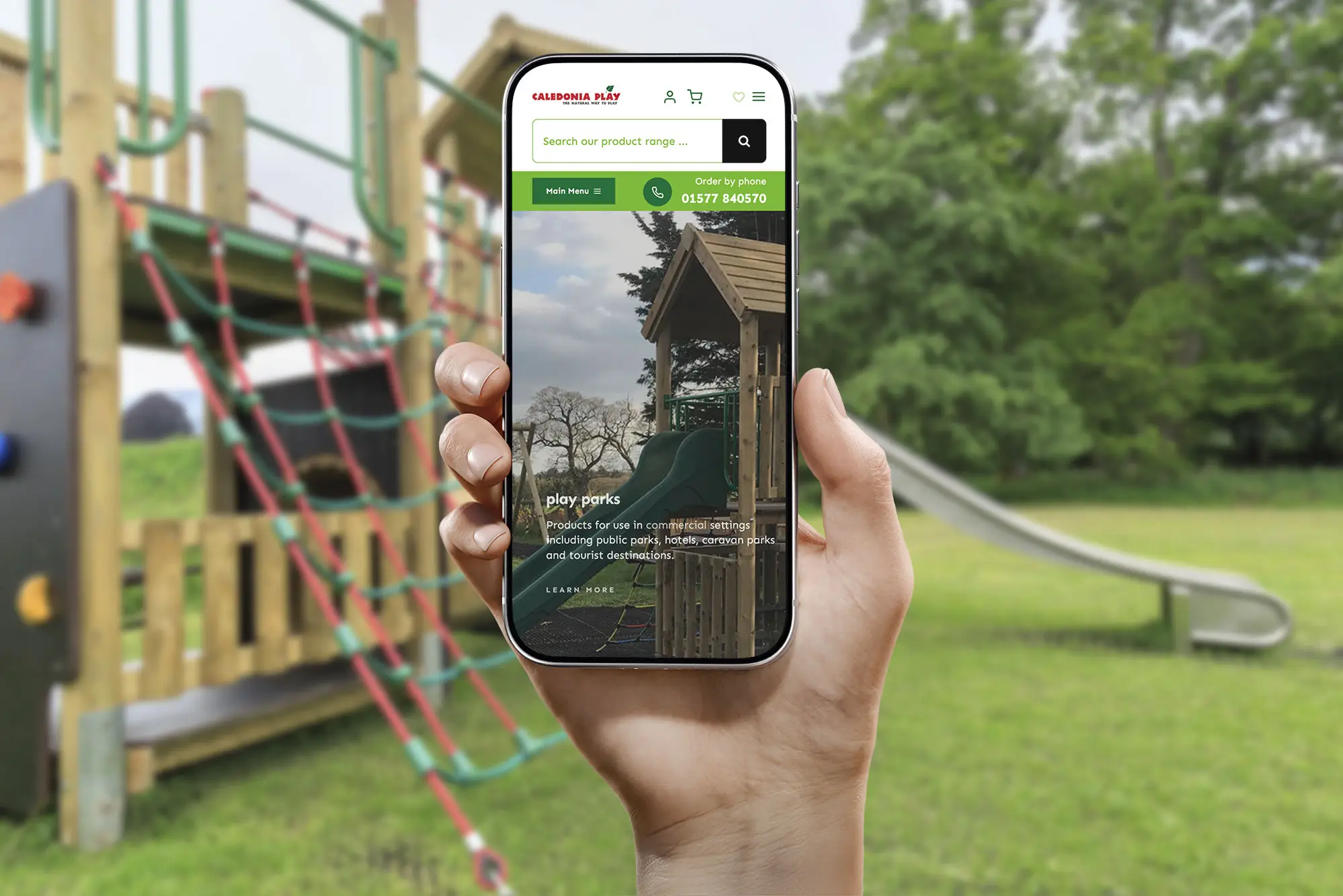 Hand holding a smartphone displaying the Caledonia Play online shop in front of playground equipment
